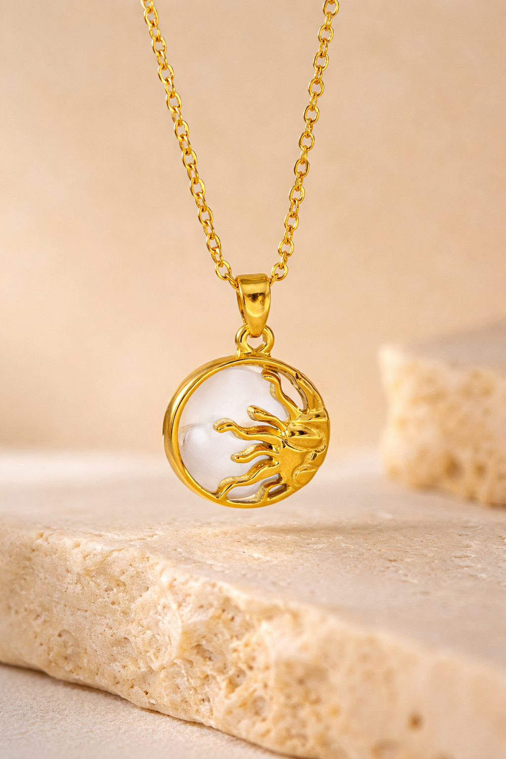Gold sun pendant necklace with white center detail displayed on a neutral stone surface, showcasing a small round charm ana a gold chain